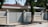 A modern house with a flat roof is surrounded by a high beige fence with metal slats and decorative spherical lights on top of the posts. It is located on a paved street with trees and greenery in the background.