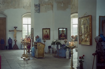 A serene church interior with several ornate religious icons and paintings on the walls. The space is minimally decorated with gold-colored candle holders and a crucifix to the left. A man is standing at a lectern, possibly in prayer or officiating, while a few people are standing quietly in the background. The lighting is soft, filtered through large windows.