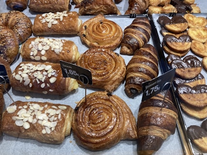 A variety of freshly baked pastries are displayed on trays, including croissants, chocolate-dipped palmiers, almond-topped pastries, and spiral cinnamon rolls. Price tags indicate the cost of some items.