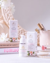 A beauty product display featuring a bottle and box of eye rescue cream. The scene is set against a neutral background with soft, warm lighting. Surrounding the product are decorative items, including white flowers, a stack of books with fashion-oriented titles, a pearl necklace, and cosmetic items, creating a luxurious and elegant atmosphere.