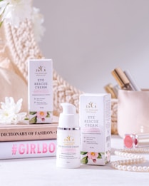 A beauty product display featuring a bottle and box of eye rescue cream. The scene is set against a neutral background with soft, warm lighting. Surrounding the product are decorative items, including white flowers, a stack of books with fashion-oriented titles, a pearl necklace, and cosmetic items, creating a luxurious and elegant atmosphere.