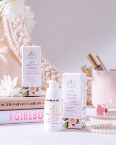 A beauty product display featuring a bottle and box of eye rescue cream. The scene is set against a neutral background with soft, warm lighting. Surrounding the product are decorative items, including white flowers, a stack of books with fashion-oriented titles, a pearl necklace, and cosmetic items, creating a luxurious and elegant atmosphere.