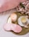 A round golden tray holds a lit candle, a teacup with saucer, a pink satin sleep mask, and a facial roller. Beside the tray are pink hydrangea flowers. The background includes a soft pink sheet.