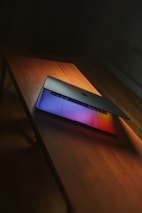 A laptop partially open on a wooden table, with vibrant colors reflecting from the screen onto the surface.