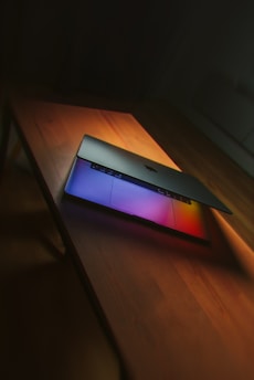 A laptop partially open on a wooden table, with vibrant colors reflecting from the screen onto the surface.