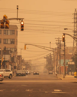 A city street is observed with a hazy atmosphere, possibly due to smog or smoke. Traffic lights hang above, showing yellow and red lights. Cars are either parked or driving along the road. Power and telephone lines crisscross the scene, contributing to urban clutter. Buildings line both sides of the street.