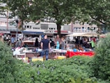 People are browsing and setting up stalls at an outdoor market. Various items such as clothes, books, and accessories are displayed on tables with vibrant tablecloths. The market is set in an urban environment, with buildings and shops in the background. A large tree provides shade, and greenery with purple flowers decorates the foreground.