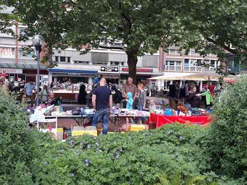 People are browsing and setting up stalls at an outdoor market. Various items such as clothes, books, and accessories are displayed on tables with vibrant tablecloths. The market is set in an urban environment, with buildings and shops in the background. A large tree provides shade, and greenery with purple flowers decorates the foreground.