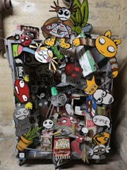 A vibrant collage of cartoon-like characters and objects attached to what seems to be a cart or container. The artwork features exaggerated facial expressions and colorful clothing. Some characters have skeletal features, and others resemble animals or playful monsters. The background is a plain wall, emphasizing the chaos and creativity of the collage.