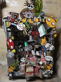 A vibrant collage of cartoon-like characters and objects attached to what seems to be a cart or container. The artwork features exaggerated facial expressions and colorful clothing. Some characters have skeletal features, and others resemble animals or playful monsters. The background is a plain wall, emphasizing the chaos and creativity of the collage.