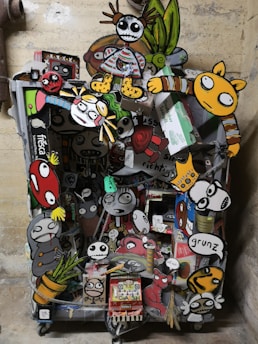 A vibrant collage of cartoon-like characters and objects attached to what seems to be a cart or container. The artwork features exaggerated facial expressions and colorful clothing. Some characters have skeletal features, and others resemble animals or playful monsters. The background is a plain wall, emphasizing the chaos and creativity of the collage.