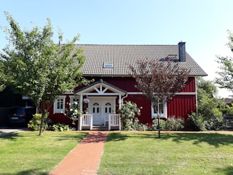 A charming red wooden house with a white-trimmed entrance and a neatly tiled roof is surrounded by a garden with green grass and trees. Brick pathway leads to the front porch. Plants and flowers adorn the area around the house, contributing to a welcoming and cozy atmosphere.