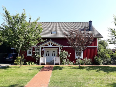 A charming red wooden house with a white-trimmed entrance and a neatly tiled roof is surrounded by a garden with green grass and trees. Brick pathway leads to the front porch. Plants and flowers adorn the area around the house, contributing to a welcoming and cozy atmosphere.