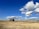 A vast, grassy field under a bright blue sky filled with fluffy white clouds. Two trees stand in the middle distance, while a pair of portable toilets is visible on the right. The area is open and stretches to an embankment in the background.