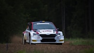 A rally car is driving on a dirt road surrounded by a forest. The car is white with red and black accents, featuring several sponsor logos. Dense green foliage forms the background, and dust is kicked up behind the vehicle as it moves swiftly.