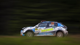 A rally car is captured in motion on a blurred background, suggesting high speed. The car is adorned with various sponsor stickers and has the number 33 prominently displayed on its side. The background is predominantly green, indicating a race through a natural or forested environment.