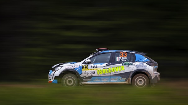 A rally car is captured in motion on a blurred background, suggesting high speed. The car is adorned with various sponsor stickers and has the number 33 prominently displayed on its side. The background is predominantly green, indicating a race through a natural or forested environment.