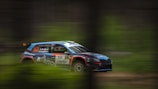 A rally car in motion is captured mid-race with a blurred background indicating speed. The car is marked with the number 52 and has various sponsor logos. It features a sporty design with a combination of blue, orange, and black colors.
