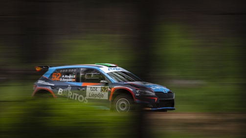 A rally car in motion is captured mid-race with a blurred background indicating speed. The car is marked with the number 52 and has various sponsor logos. It features a sporty design with a combination of blue, orange, and black colors.