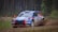 A rally car with colorful sponsorship decals races on a dirt path through a forest. The car's dynamic motion is captured as it kicks up dust and gravel from the road. Tall trees line the background, and the setting suggests a competitive motorsport event.