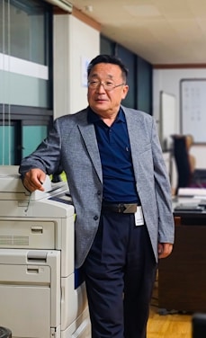 An older man wearing glasses and a gray blazer is standing next to a copier machine in what appears to be an office environment. He has one hand resting on the copier and is looking away from the camera, giving a relaxed and contemplative impression. The office space features a window with blinds, office furniture, and equipment in the background.
