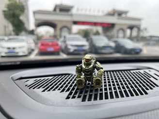 A small, gold-colored toy robot sits on a car dashboard with a parking lot and an entrance gate with an archway visible outside the windshield. The focus is on the toy while the background is slightly blurred, featuring several parked cars.