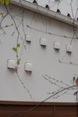 A series of white ventilation covers or fixtures are mounted on an exterior wall. The wall is made of light-colored material, and there are branches with sparse leaves in the foreground.