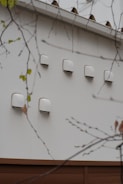 A series of white ventilation covers or fixtures are mounted on an exterior wall. The wall is made of light-colored material, and there are branches with sparse leaves in the foreground.