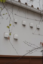 A series of white ventilation covers or fixtures are mounted on an exterior wall. The wall is made of light-colored material, and there are branches with sparse leaves in the foreground.