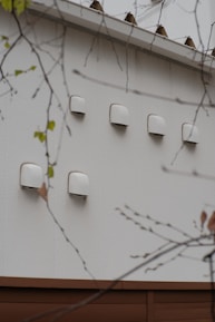 A series of white ventilation covers or fixtures are mounted on an exterior wall. The wall is made of light-colored material, and there are branches with sparse leaves in the foreground.