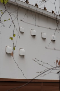 A series of white ventilation covers or fixtures are mounted on an exterior wall. The wall is made of light-colored material, and there are branches with sparse leaves in the foreground.