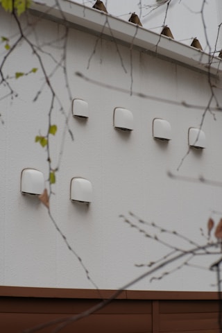A series of white ventilation covers or fixtures are mounted on an exterior wall. The wall is made of light-colored material, and there are branches with sparse leaves in the foreground.