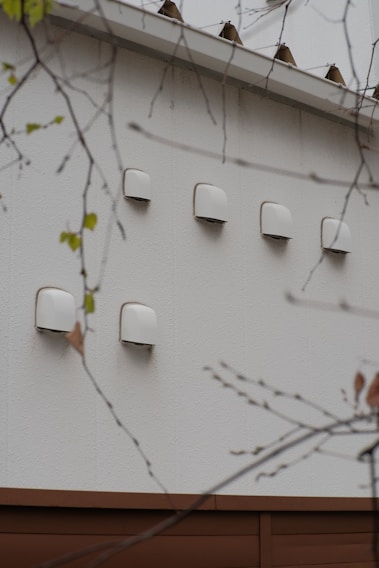 A series of white ventilation covers or fixtures are mounted on an exterior wall. The wall is made of light-colored material, and there are branches with sparse leaves in the foreground.