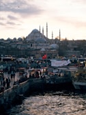 A bustling waterfront scene with numerous people gathered near stalls and booths. In the background, a large historic mosque with multiple domes and minarets dominates the skyline. Several Turkish flags are visible, and the sun is setting, casting a warm glow over the entire scene. The water in the foreground reflects the surroundings.