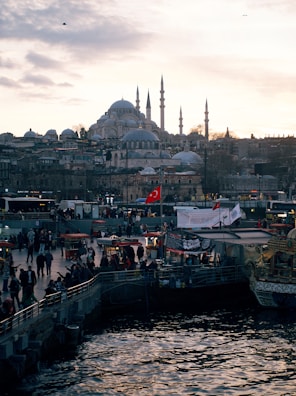 A bustling waterfront scene with numerous people gathered near stalls and booths. In the background, a large historic mosque with multiple domes and minarets dominates the skyline. Several Turkish flags are visible, and the sun is setting, casting a warm glow over the entire scene. The water in the foreground reflects the surroundings.