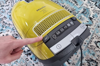 A yellow vacuum cleaner with a control panel featuring various icons for settings. A finger is pressing a button on the panel. The vacuum is placed on a patterned carpet with shades of blue, white, and black.
