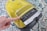 A yellow vacuum cleaner with a control panel featuring various icons for settings. A finger is pressing a button on the panel. The vacuum is placed on a patterned carpet with shades of blue, white, and black.