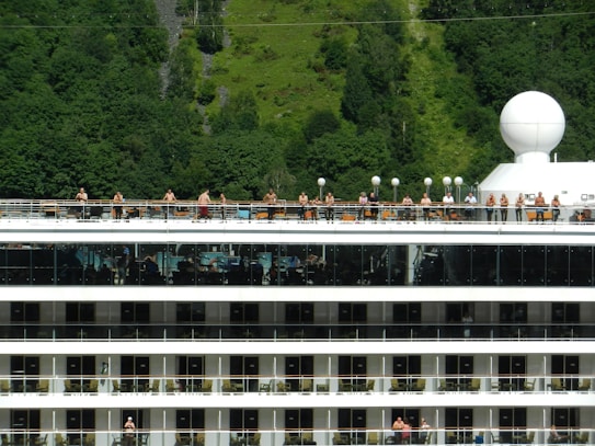 A large cruise ship with multiple decks is shown, featuring people standing on one of the upper decks. The background consists of dense, green forested hills. The top of the ship has circular white structures, and the decks have reflective glass and railings.