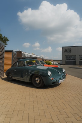 A classic vintage car with sleek, rounded lines and a glossy dark green finish is parked on a paved area. In the background, a modern industrial building labeled A3 can be seen under a bright sky filled with fluffy clouds. The scene is set in an urban area with some greenery visible to the side.