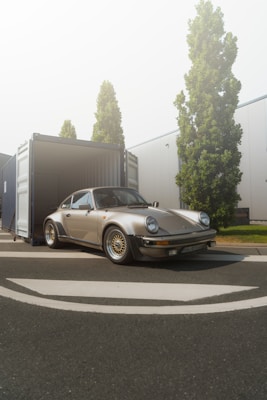 A classic sports car is parked partially outside an open shipping container in an industrial area. Tall green trees line the background alongside white industrial buildings. The road is clear and the sun casts a gentle light, creating a calm atmosphere.