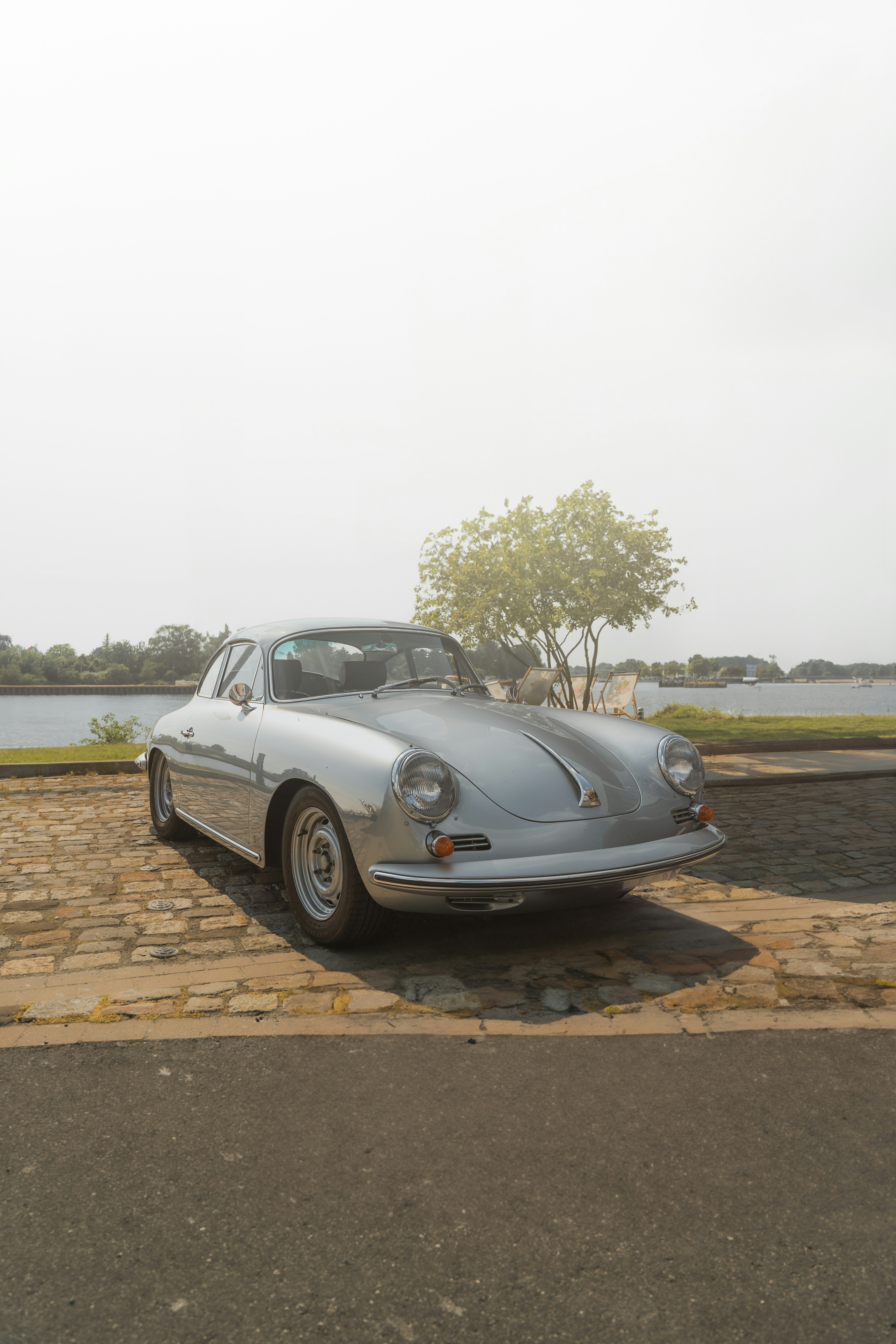 Auto sportiva d'epoca argentata parcheggiata sui ciottoli vicino a un lago.