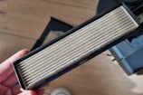A rectangular air filter is held in a person's hand. The filter has visible white pleats and is encased in a black frame. It appears to be new or unused. The background includes a wooden floor and part of a device or machine.