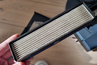 A rectangular air filter is held in a person's hand. The filter has visible white pleats and is encased in a black frame. It appears to be new or unused. The background includes a wooden floor and part of a device or machine.