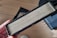 A rectangular air filter is held in a person's hand. The filter has visible white pleats and is encased in a black frame. It appears to be new or unused. The background includes a wooden floor and part of a device or machine.