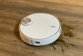 A white robotic vacuum cleaner is on a wooden floor, next to a spilled liquid that appears to be brown. The robot has a round design with a metallic disc on top and buttons with blue lights.