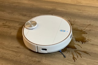 A white robotic vacuum cleaner is on a wooden floor, next to a spilled liquid that appears to be brown. The robot has a round design with a metallic disc on top and buttons with blue lights.