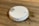 A white robotic vacuum cleaner is on a wooden floor, next to a spilled liquid that appears to be brown. The robot has a round design with a metallic disc on top and buttons with blue lights.