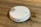 A white robotic vacuum cleaner is on a wooden floor, next to a spilled liquid that appears to be brown. The robot has a round design with a metallic disc on top and buttons with blue lights.