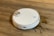 A white robotic vacuum cleaner is on a wooden floor, next to a spilled liquid that appears to be brown. The robot has a round design with a metallic disc on top and buttons with blue lights.