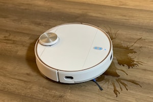 A white robotic vacuum cleaner is on a wooden floor, next to a spilled liquid that appears to be brown. The robot has a round design with a metallic disc on top and buttons with blue lights.